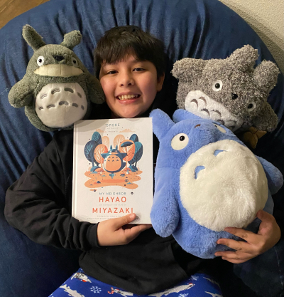 A boy laying down on a bean bag chair holding a book with 3 Totoro toys around him